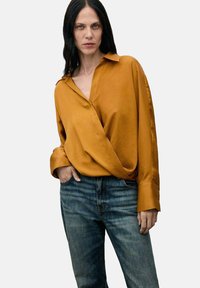 Camisa envolvente de color mostaza en tejido suave con cuello; manga larga, con puños de botón. Combinada con jeans de mezclilla azul, con un leve desgaste.