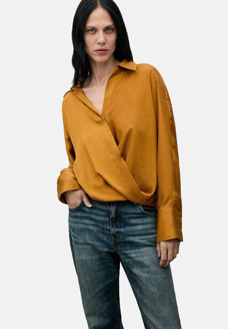 Camisa envolvente de color mostaza en tejido suave con cuello; manga larga, con puños de botón. Combinada con jeans de mezclilla azul, con un leve desgaste.