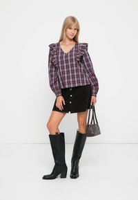 Geruite blouse met ruches aan de mouwen, zwarte corduroy rok met knopen aan de voorkant, kniehoge zwarte laarzen en een kleine grijze handtas.