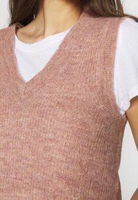 Junge Person, die eine weiche, rosa gestrickte ärmellose Weste über einem weißen T-Shirt mit kurzen Ärmeln trägt, sichtbar von den Schultern bis zur Brust.