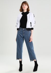 Vit bomberjacka med svarta detaljer, kombinerad med högmidjade blå vida jeans. Modellen bär svarta ankelskor och en åtsittande svart topp.