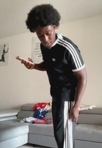 Νέος άνδρας με μαύρη φόρμα Adidas ρυθμίζει το παντελόνι κρατώντας ένα smartphone σε σαλόνι με διπλωμένα ρούχα στον καναπέ.