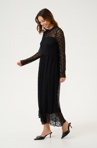 Rochie din dantelă neagră cu mânecă lungă, decolteu înalt și o siluetă fluidă. Pantofii sunt negri, cu vârfuri ascuțite și accente decorative.