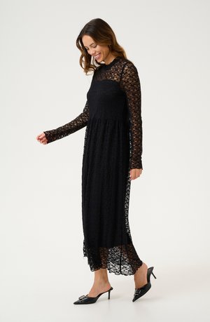 Woman smiling, wearing a long black lace dress with sheer sleeves and black pointed-toe heeled mules, standing on a white background.