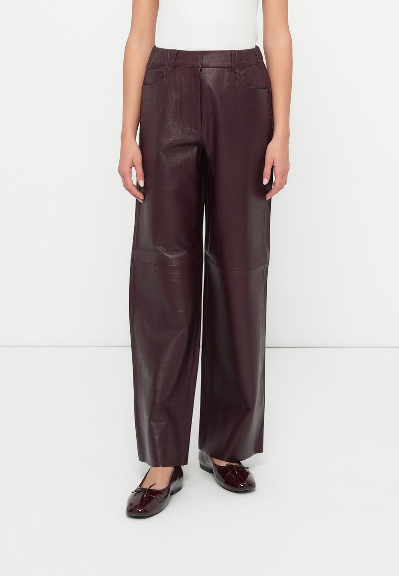 Vrouw in hoge taille, wijde bordeaux faux leren broek gecombineerd met bijpassende glanzende ballerina's, staand op een witte vloer.