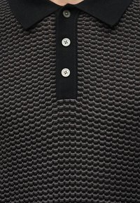 Μαύρο πουλόβερ με γιακά, με υφή κυματιστού μοτίβου και τρία λευκά κουμπιά στην πατιλέτα.