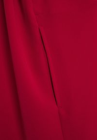 Tissu rouge à la texture lisse, avec une poche latérale. Des coutures sont visibles le long du bord de la poche pour plus de détail.