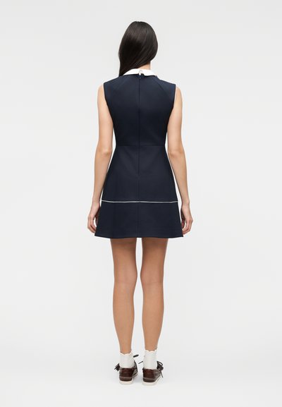 Γυναίκα που στέκεται με την πλάτη, φορώντας αμάνικο σκούρο μπλε φόρεμα με λευκές λεπτομέρειες, συνδυασμένο με λευκές κάλτσες και καφέ παπούτσια με κορδόνια.