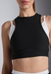 Musta ribbed crop top valkoisilla olkaimilla, jossa on korkea kaula-aukko ja ommeltu yksityiskohta keskiosassa. Sileä pinta ja mukautuva muotoilu.