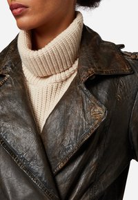 Brun läderjacka med en sliten textur och överdimensionerad krage, lager-på-lager över en krämfärgad ribbad polotröja.