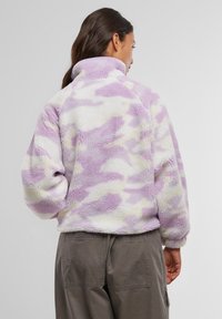 Fialová a krémová fleecová bunda s mäkkým, textúrovaným camouflage vzorom, vysokým golierom a elastickými manžetami. Nosená cez olivovo zelené nohavice.