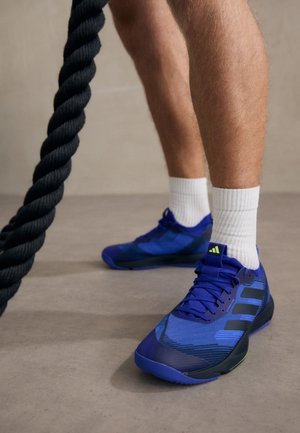 Personne portant des chaussures de sport bleues et des chaussettes blanches, tenant une corde de combat épaisse noire sur un sol de salle de sport.