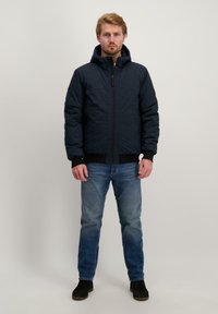 Marineblauwe pufferjack met een capuchon, met een ritssluiting aan de voorkant en geribbelde manchetten. Gecombineerd met een blauwe spijkerbroek en zwarte enkellaarsjes.