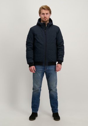 Marineblauer Puffer-Jacke mit Kapuze, ausgestattet mit einem Frontreißverschluss und gerippten Bündchen. Kombiniert mit blauen Jeans und schwarzen Stiefeletten.