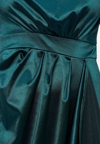 Γυαλιστερό σκούρο πράσινο σατέν ύφασμα με πιετά και φαρδιά ζώνη στη μέση, δημιουργώντας ομαλές και υφασματικές πτυχές.