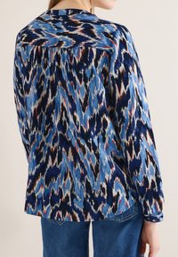 Femme portant un chemisier à manches longues avec un motif abstrait bleu, noir, blanc et marron, assorti à un jean bleu, vue de dos.