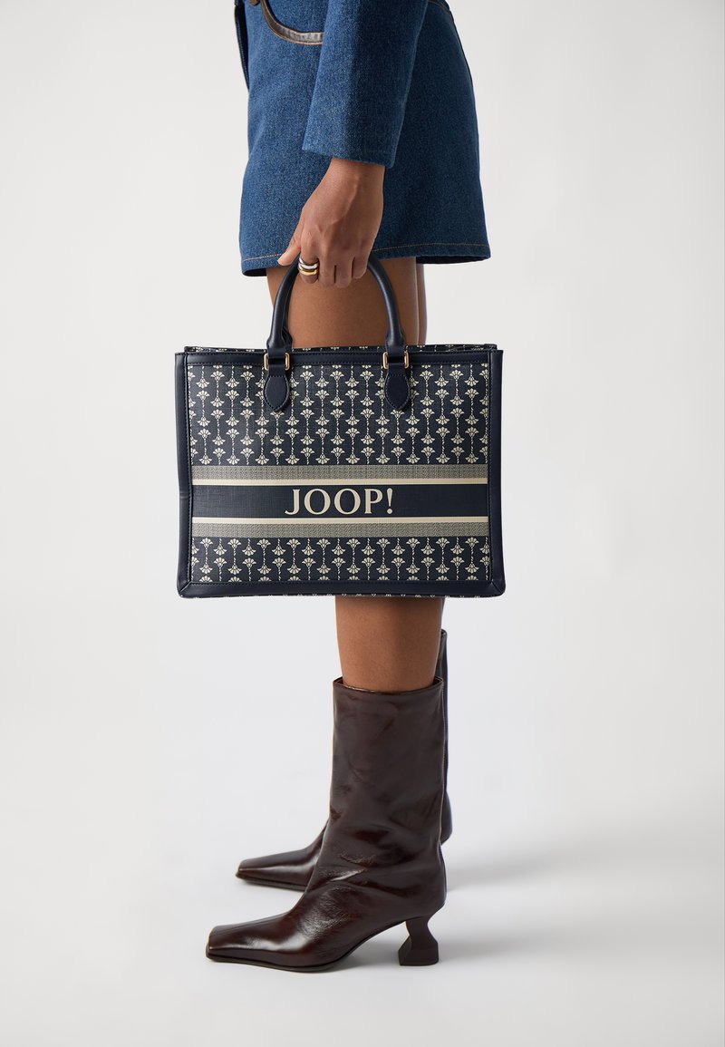Tamnoplava torba s ponavljajućim cvjetnim uzorkom, naglašena bež prugom s "JOOP!" logotipom. Glatka tekstura s dva ručka.