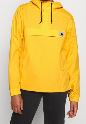 Žlutá bunda s kapucí, černými stahovacími šňůrkami, klopou na přední kapse a malou značkovou nášivkou na hrudi, nošená s černými kalhotami.