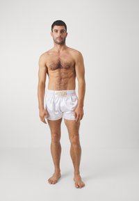 White satin shorts with a gold logo waistband, featuring a loose fit and smooth texture, worn by a person standing barefoot.
