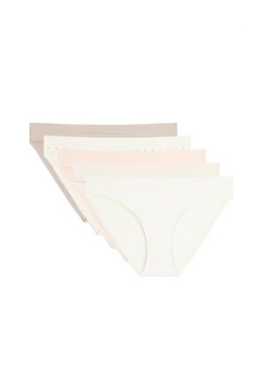 Femme portant un sous-vêtement beige sans coutures à taille basse, debout devant un fond clair uni.