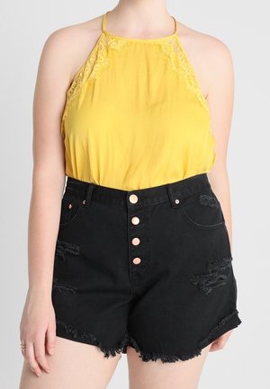 Nainen pukeutuneena hihattomaan keltaiseen haltertoppiin, jossa on pitsiyksityiskohtia, yhdistettynä korkeavyötäröisiin mustiin kulutettuihin farkkushortseihin, joissa on nappikiinnitys.