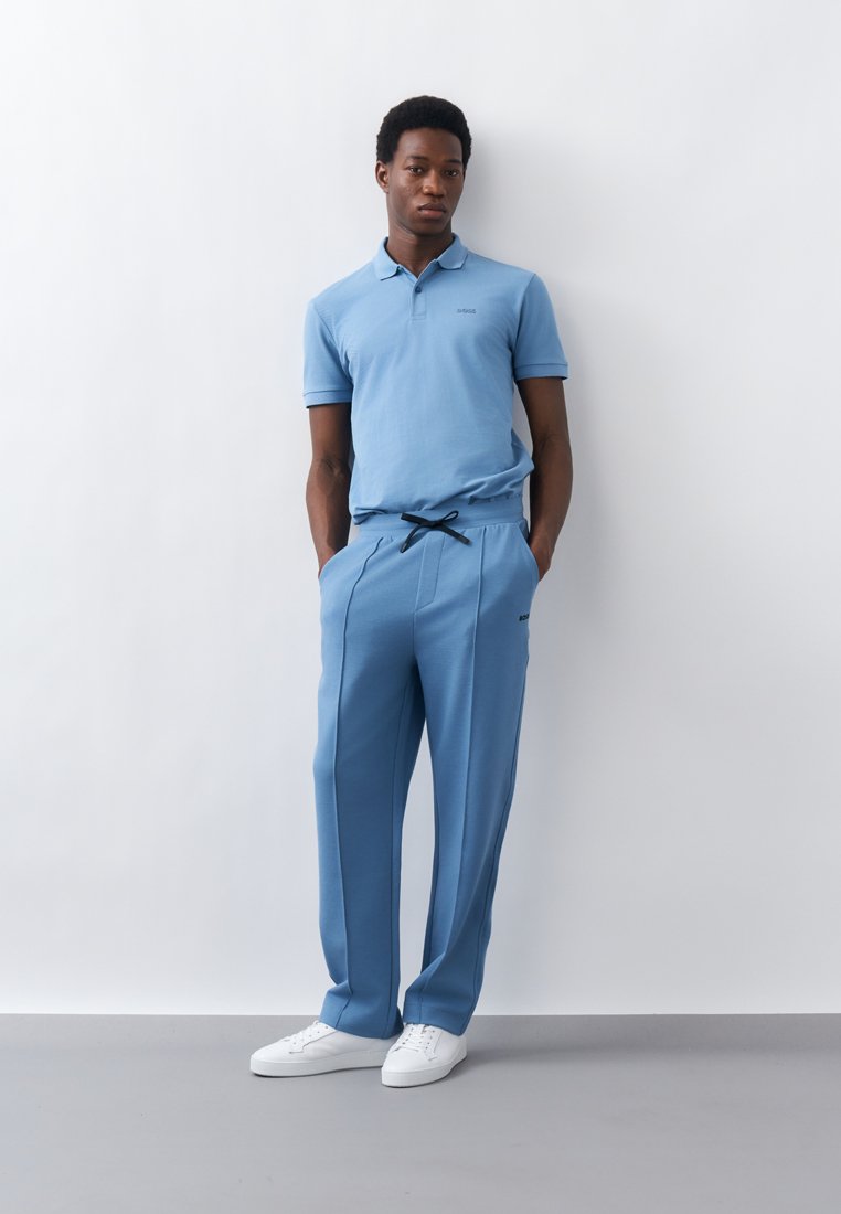 Homme debout contre un mur blanc, portant un polo bleu clair, un pantalon assorti avec un cordon noir, et des baskets blanches, les mains dans les poches.