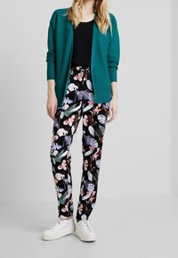 Cardigan teal con texture a coste, t-shirt nera e pantaloni neri con stampa floreale che presenta foglie e fiori in rosa e blu. Sneakers bianche.