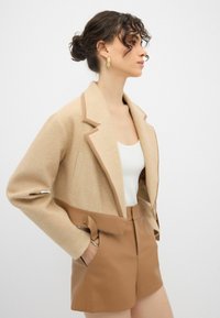 Beige cropped jakke med lysebrun faux læder taljebånd, med en notched krave og sidelommer, parret med matchende brune shorts.