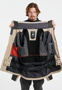 Jas met een beige buitenkant en donkergrijze voering, met oranje accenten en mesh vakken. Labels aan de binnenkant geven merkdetails aan.