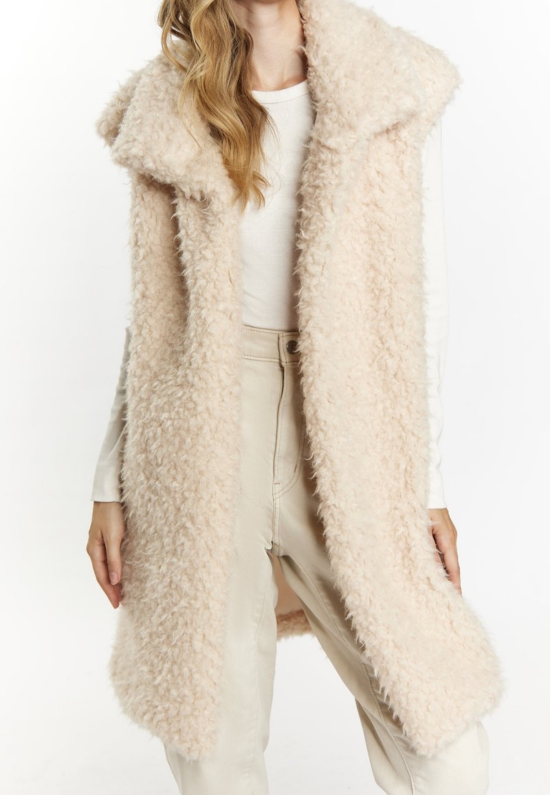 Femme portant une longue veste beige duveteuse sans manches sur une chemise blanche à manches longues et un pantalon beige clair, vue du cou jusqu'à mi-cuisse.