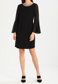 Robe noire avec des manches cloche transparentes et un col en perles. Forme A, longueur genoux, texture lisse, portée avec des chaussures noires à talons pointus.