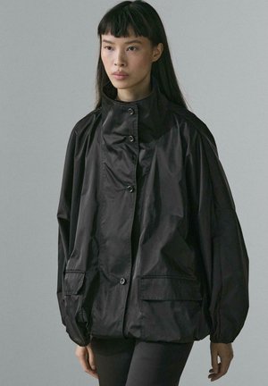 Giovane donna che indossa una giacca nera oversize con bottoni, colletto alto e grandi tasche anteriori su uno sfondo grigio uniforme.