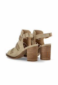 Botas de tobillo de ante beige con diseño abierto, un tacón curvado y detalles trenzados. Incluyen una correa con hebilla y un tacón de bloque a rayas.