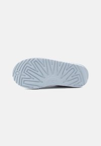 Gaiši zils gumijas apavu zole ar teksturētu dizainu, kurā ir zigzaga raksti un iegravēts "UGG" logo tuvu papēdim.