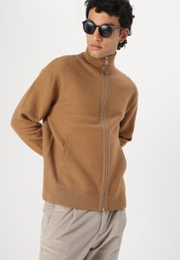 Brun zip-up genser laget av myk stoff med høy krage, med sidelommer og ribbestrikkede mansjetter og hem. Modell iført runde solbriller.