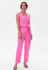 Roze linnen twee-delige outfit met een mouwloze vest met drie knopen en hoge taillebroeken met een gestrikte taille. Aangevuld met hakken.