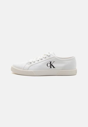 Hvit lerrets sneakers med lav toppdesign, med gummisåle og svart "CK"-logo på siden. Seks hull med hvite lisser.