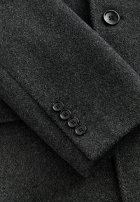 Grå ullkappa ärm med texturerat tyg, med fyra svarta knappar vid manschetten. Kappan har en skräddarsydd design.