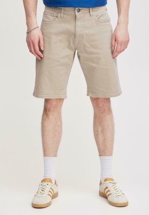 Uomo che indossa pantaloncini beige al ginocchio, calzini bianchi e scarpe da ginnastica bianche con strisce marroni, in piedi contro uno sfondo chiaro e semplice.