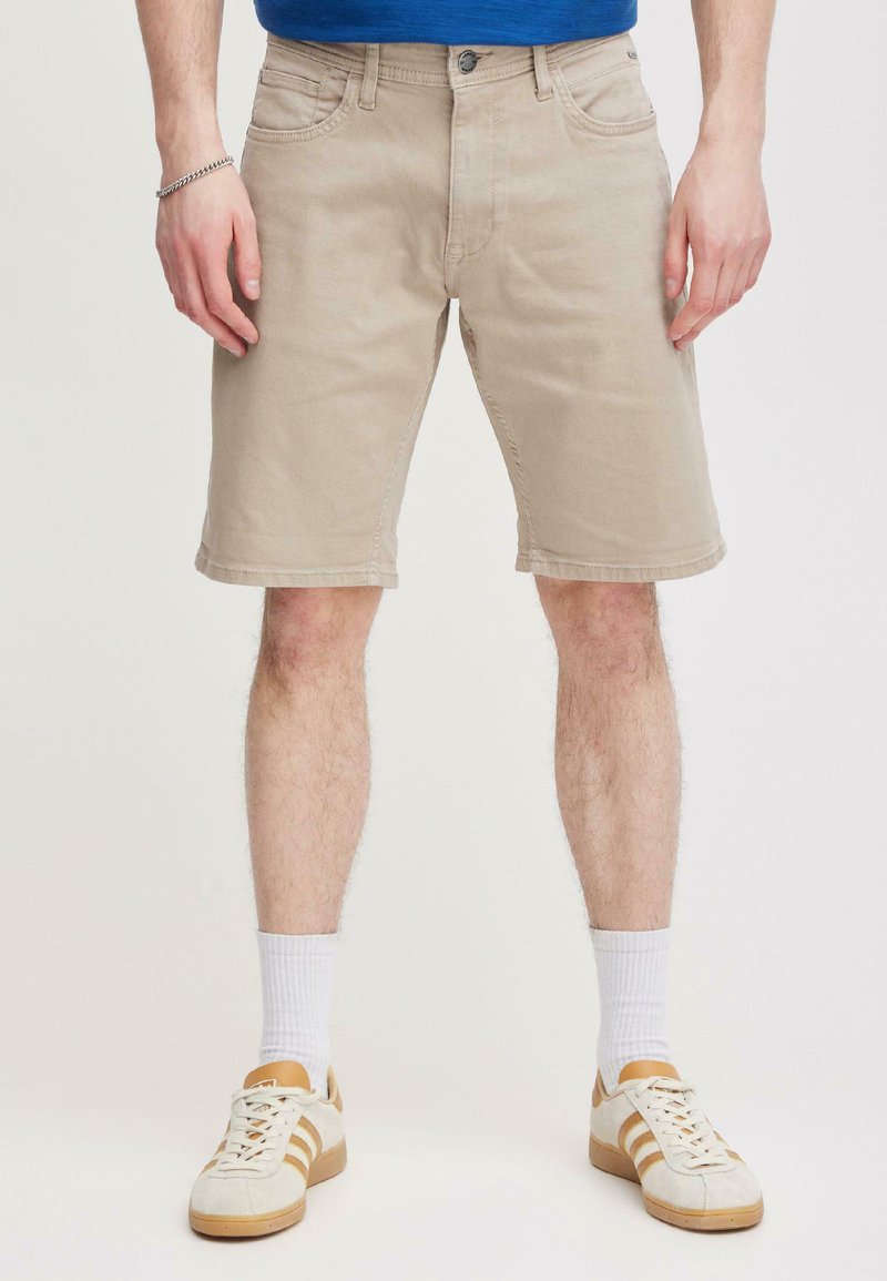 Homme portant un short beige au niveau des genoux, des chaussettes blanches et des baskets blanches à rayures marron, debout devant un fond clair et uni.