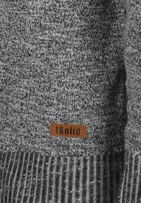 Pull en laine grise avec un motif texturé. Poignets et ourlet côtelés avec une étiquette en tissu beige portant l'inscription "!Solid" cousue à l'avant.