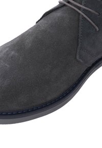 Stivaletto in suede grigio con punta tonda, lacci sottili e cucitura blu lungo la suola. Presenta una superficie testurizzata e una suola in gomma flessibile.
