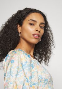 Blusa com padrão floral e mangas bufantes, nas cores azul pastel, amarelo e rosa. Feita de tecido leve com detalhes franzidos.