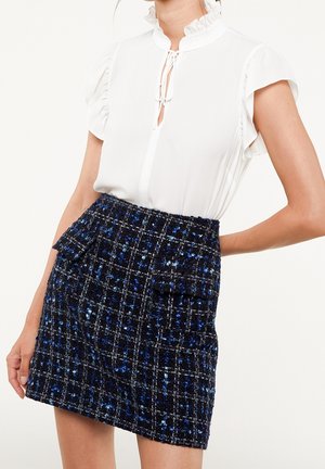 Vrouw draagt een witte mouwloze blouse met gerimpelde kraag en strik, gecombineerd met een blauw-zwarte geruite textuur mini-rok met voorzakken.