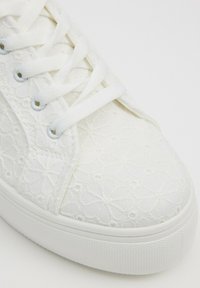 Zapatillas blancas con cordones que presentan un patrón floral texturizado, material de lona y suela de goma con borde elevado. Diseño ligero y transpirable.