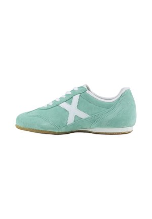Zapatilla deportiva de ante verde claro con cordones blancos, diseño blanco "X" en el lateral y suela plana de goma sobre fondo blanco.