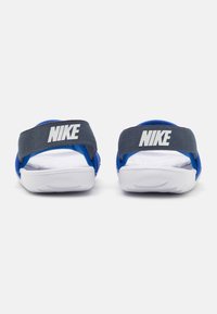 Siniset Nike-sandaalit valkoisilla pohjilla, joissa on harmaa hihna Nike-logolla. Suunnittelu on avonainen varpaista, ja siinä on pehmustettu pohja mukavuutta varten.