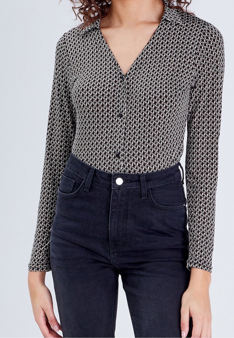 Femme portant une chemise noire et blanche à manches longues à motifs, rentrée dans un jean noir taille haute.