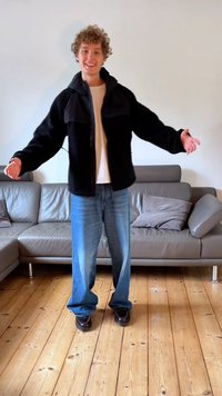 Giacca nera con zip dal tessuto morbido, indossata sopra una maglietta bianca; jeans blu larghi e scarpe scure completano l'outfit.