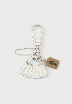 Llavero plateado con un colgante de metal en forma de abanico y una pequeña etiqueta color beige claro con el logotipo de un cocodrilo verde, sujeto a una cadena de bolas y un clip.